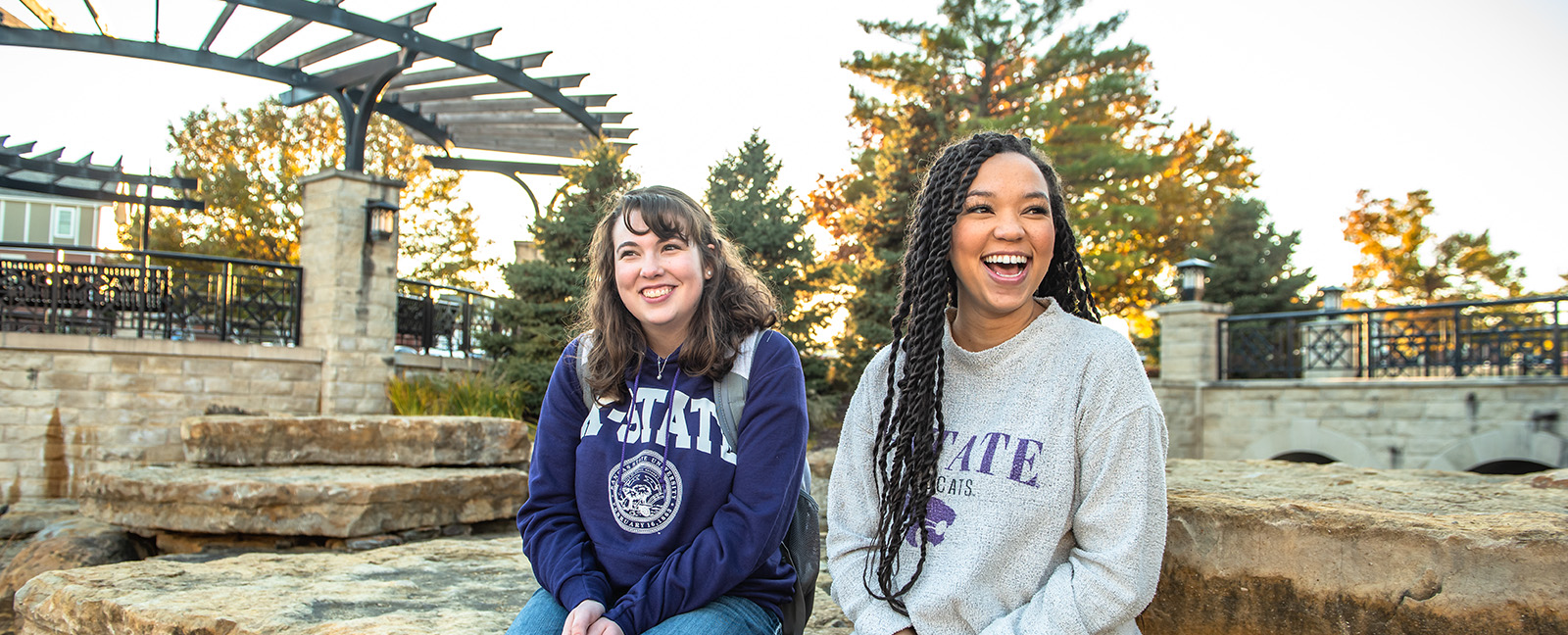 Two students sit near jardine, smiling and looking into the distance, they are wearing K-state sweatshirts