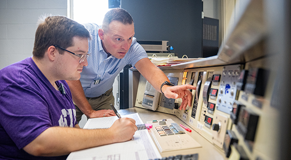 A college professor shows a student a dial on a nuclear reactor control panel.