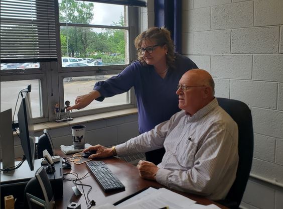 A woman gives guidance to a man sitting at a computer. 