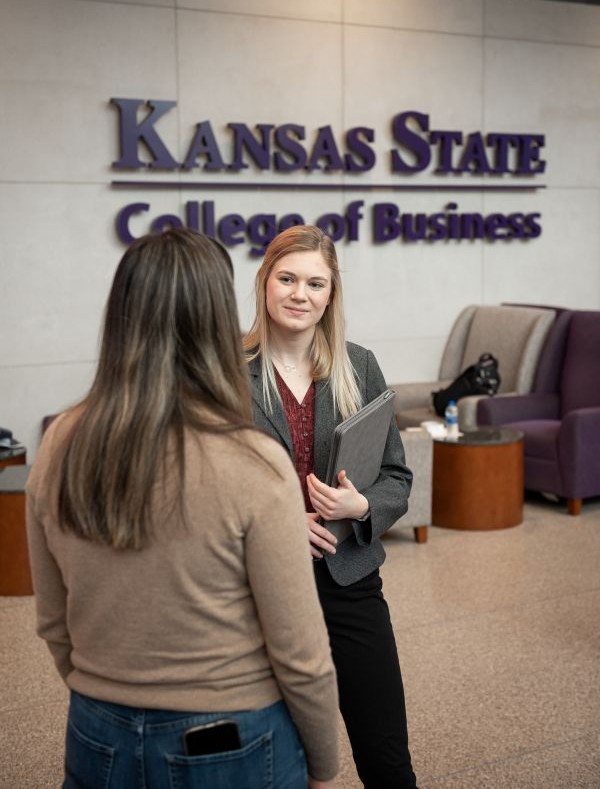 Two women converse in front of the Kansas State College of Business interior sign.