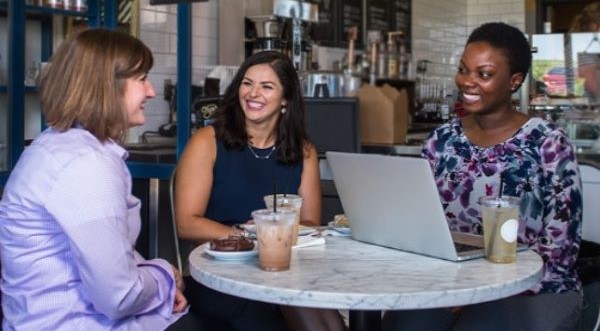 Three femals sitting at a cafe table with a laptop and a discussion happening.