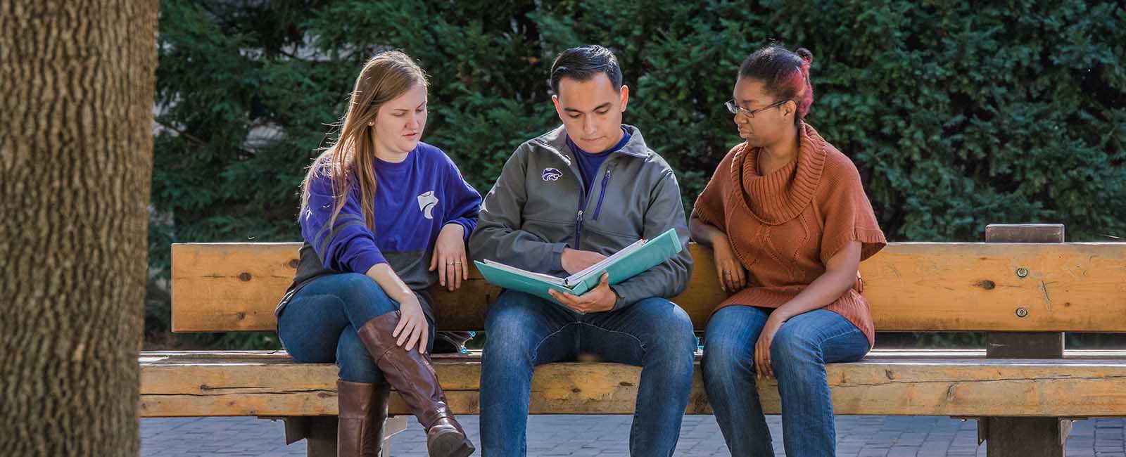 Three students (two female, one male) sitting on a bench reading a textbook. The male is in the center holding the book. The females are on either side of him, looking at the book.