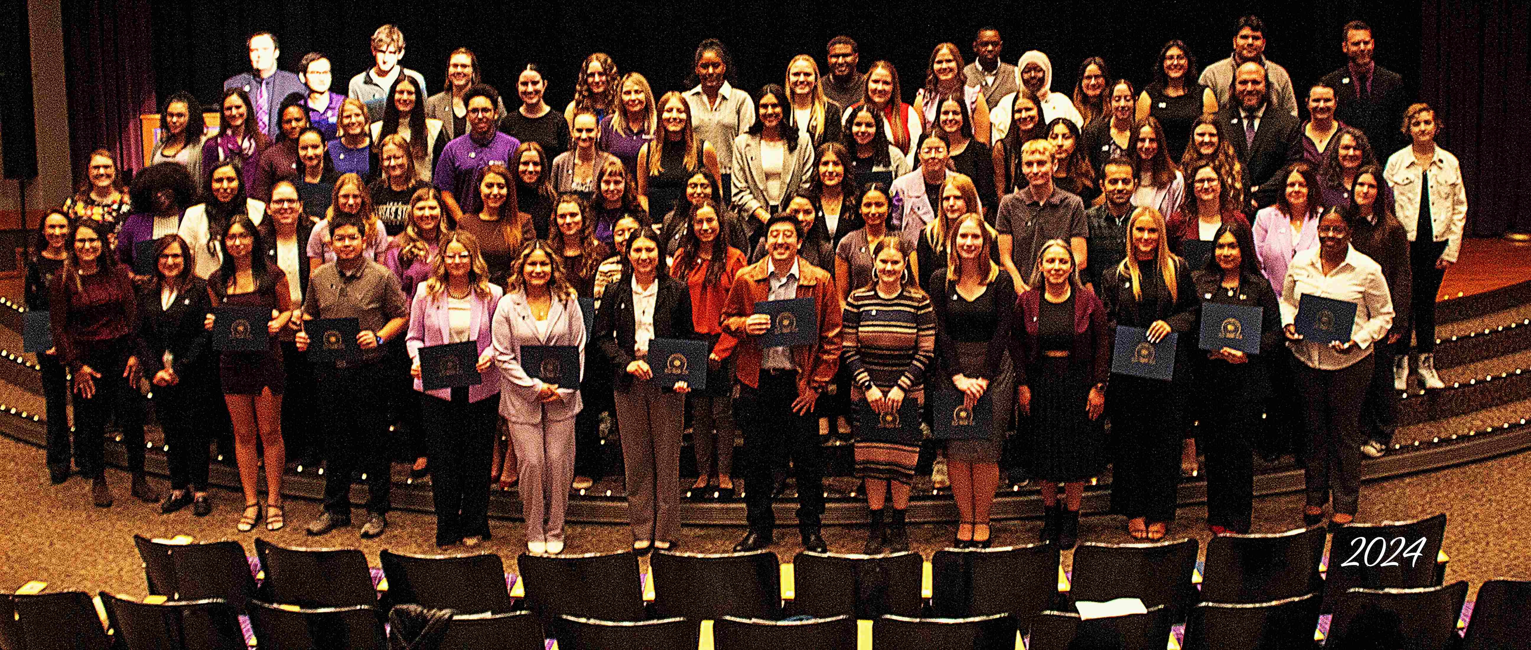 Large group of students standing on auditorium stairs holding Tri-Alpha certificates.