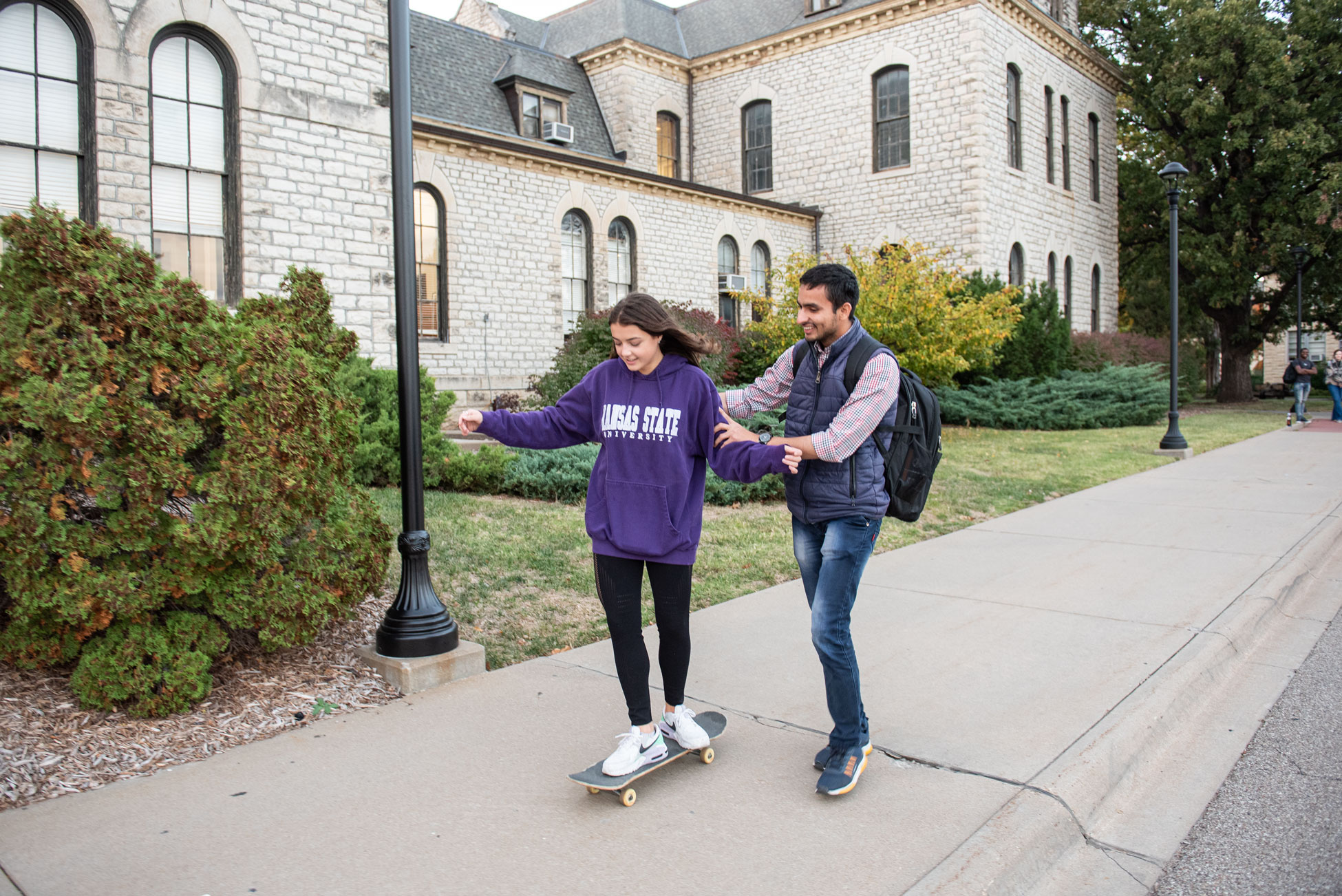 K-State student helping another student ride a skateboard on campus