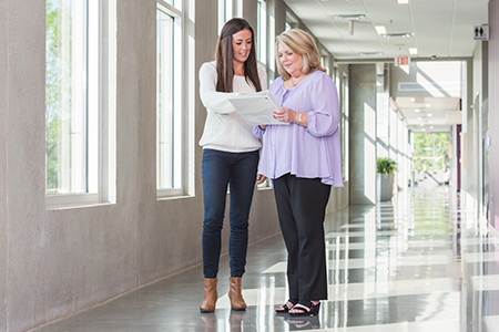 two women walking down a hallway looking at a sheet of paper held between them