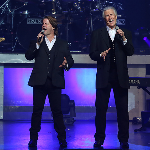 2-white-men-in dark-suits-on-stage-singing-and-holding-microphones