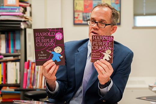 Sitting at a cluttered desk, Kansas State University children's literature professor Philip Nel holds up a modern and antique copy of "Harold and the Purple Crayon."