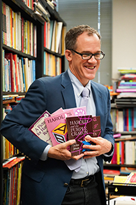 Wearing a navy suit, Philip Nel stands in his office, in front of a packed bookshelf full of colorful children's books, while he holds several international translations of children's literature classic "Harold and the Purple Crayon."
