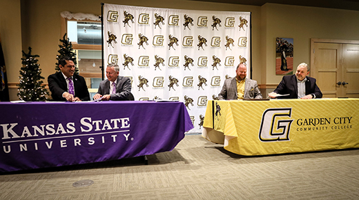 On the left, two men sit behind a table with a purple tablecloth saying "Kansas State University", while on the right, two men sit behind a different table with a yellow cloth that says "Garden City Community College."