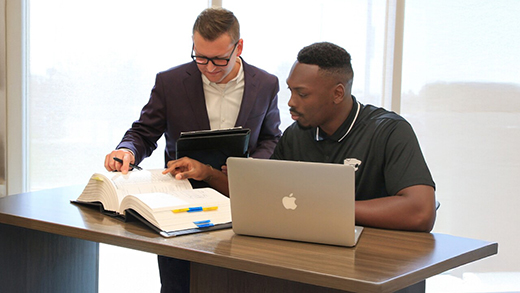 A professor stands over and assists a student writing at a laptop.