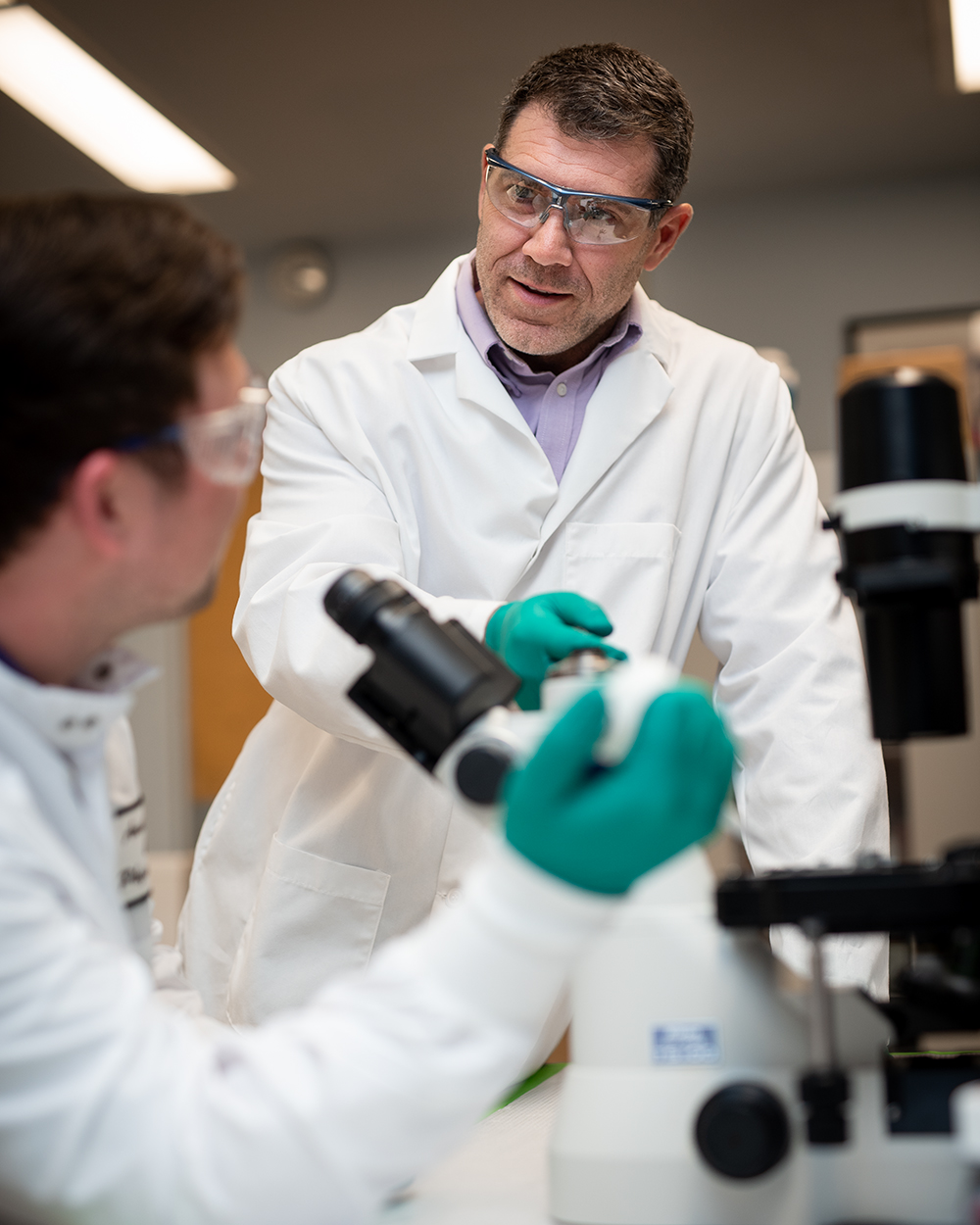 A professor in a lab coat watches as a student, also in a lab coat, uses a microscope in a laboratory.