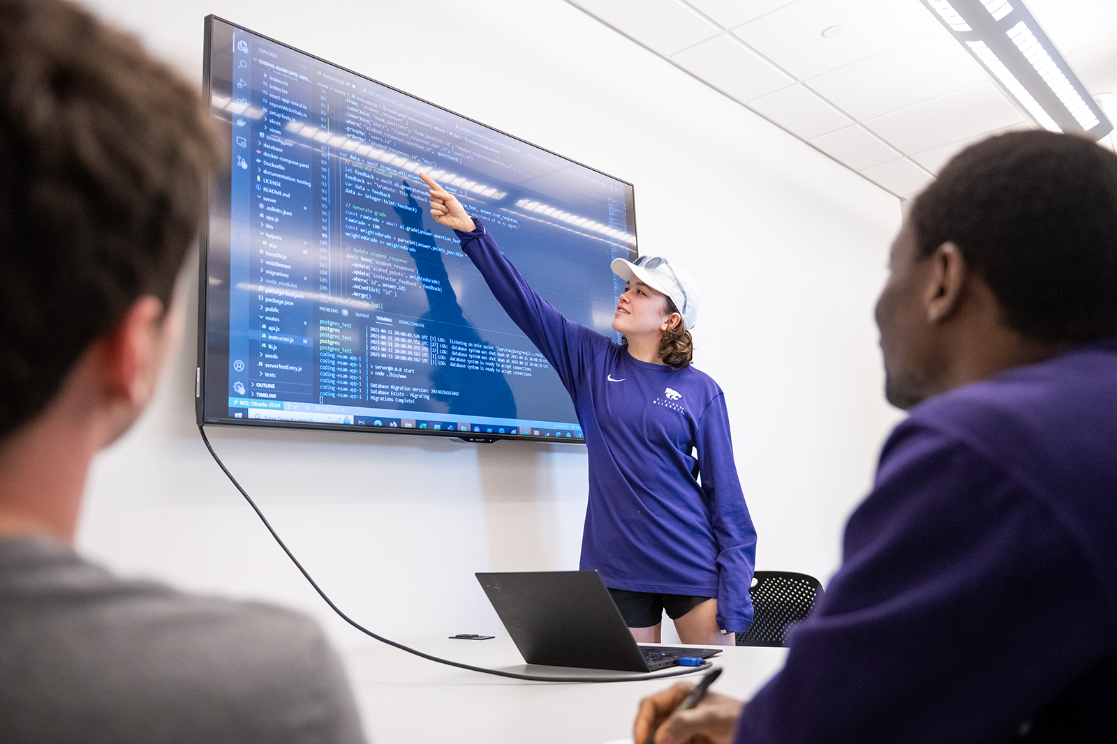 A student points at a television hanging on a wall, with several pieces of code displayed.