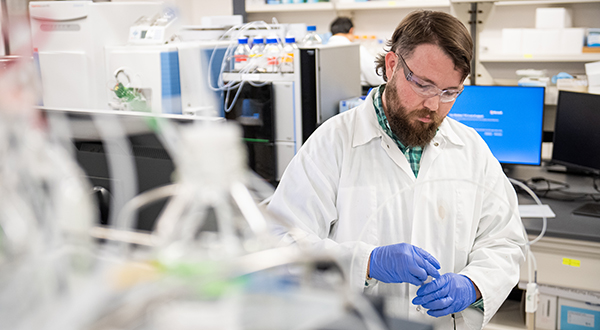 A researcher in a white lab coat and blue latex gloves works at a laboratory bench.