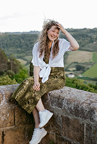 A young woman sitting on a ledge with a scenic background of green trees and fields.