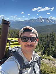 A selfie of a young man outdoors with a mountain behind him.