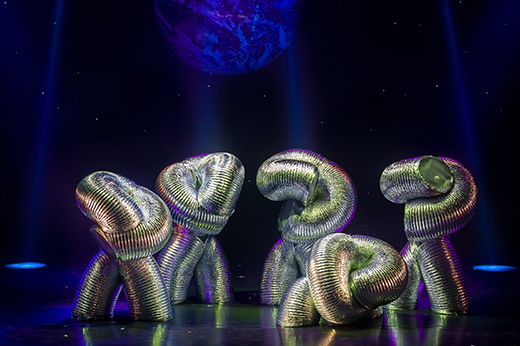 Performers dressed as aluminum tubing pose on a dark stage.