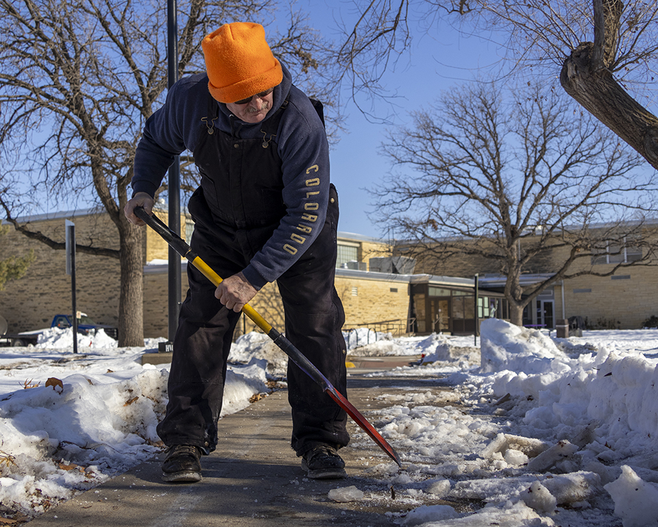 A worker in dark overalls and an orange beanie uses a sharp shooter spade to pick ice off of a university sidewalk.