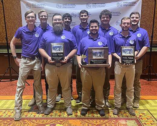 Various college-aged students, all men and in purple polos and khaki pants, stand in front of a banner holding awards they won at an international quarter-scale tractor design competition.