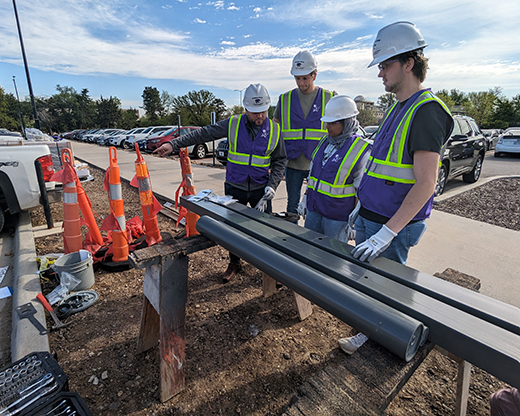 Derek Clements, assistant professor of architectural engineering and construction science, overlooks engineering plans with a group of students as they install the metal column and base for a solar-powered, off-grid EV charging station in parking lot A28, also known as the engineering parking lot. 