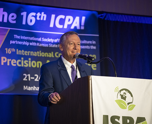 Wearing a navy suit and purple tie, Kansas State University President Richard Linton smiles as he addresses attendees inside a convention ballroom at the International Conference on Precision Agriculture.