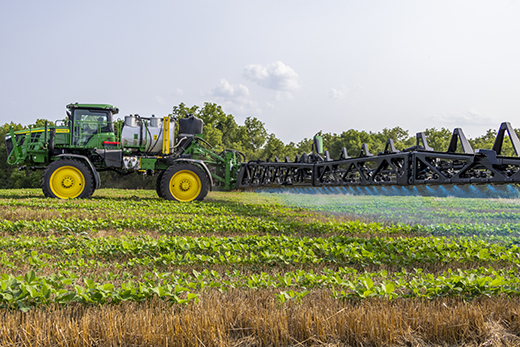 A green tractor pulls a long array of smart sprayers that precisely target weeds with a blue-tinted pesticide.