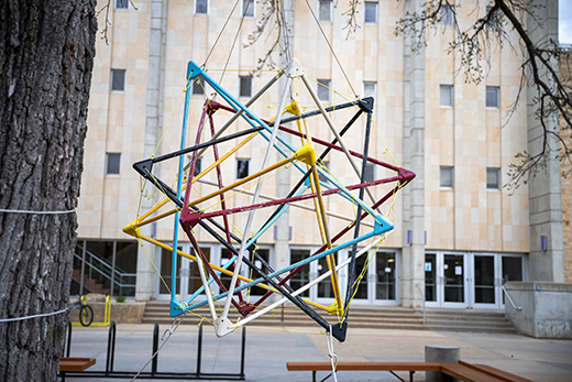 A large spherical sculpture, made up of variously colored triangle tube frames, hangs on a tree in front of the large, square limestone facade of Cardwell Hall on K-State's campus.