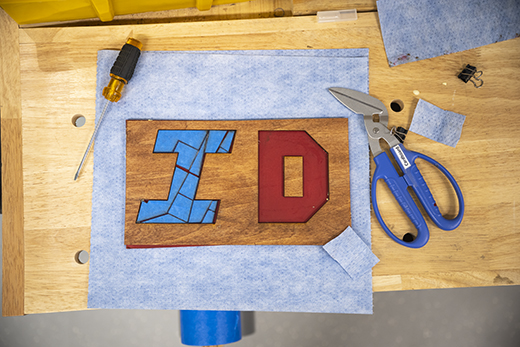 A top-down view on piece of light blue fabric on a wooden counter shows two letters — a blue I and a red D — which themselves are made up of smaller angular shapes. Tools such as a yellow screwdriver and angled, blue clippers surround the letter shapes.