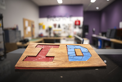 An angled view on piece of light blue fabric on a wooden counter shows two letters — a blue I and a red D — which themselves are made up of smaller angular shapes. Tools such as a yellow screwdriver and angled, blue clippers surround the letter shapes.