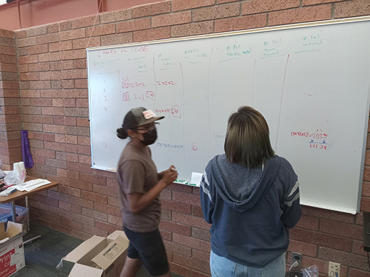 Two girls, one wearing a black face mask, work on several columns of math problems involving geometry and the numbers of sides on cubes, all written in marker on a white board hung up on a red brick wall.