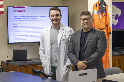 Britton Scheurmann, wearing a white lab coat, and Carl Ade, in a gray sport coat, stand in a laboratory space, with a television behind them displaying a paper that Scheuermann wrote and published in the JAMA Open Network academic journal.