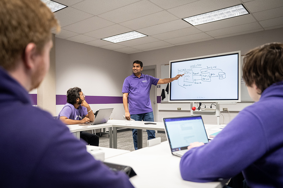 A professor in a purple polo stands in front of an interactive display board in a classroom, with students sitting in tables around the classroom.