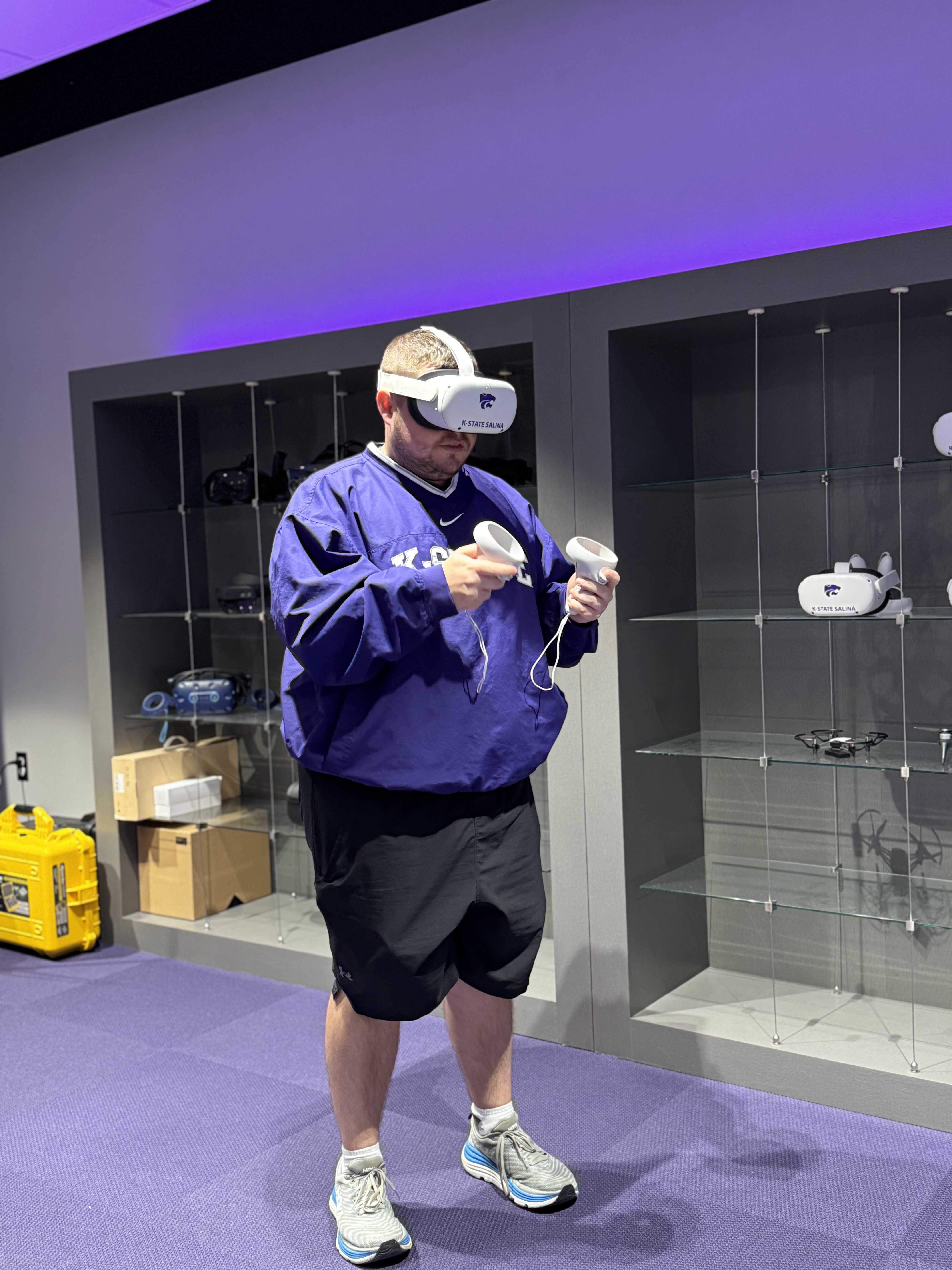 A student in a purple hoodie stands and uses a white virtual reality headset, as well as handheld paddles used to manipulate the virtual environment.