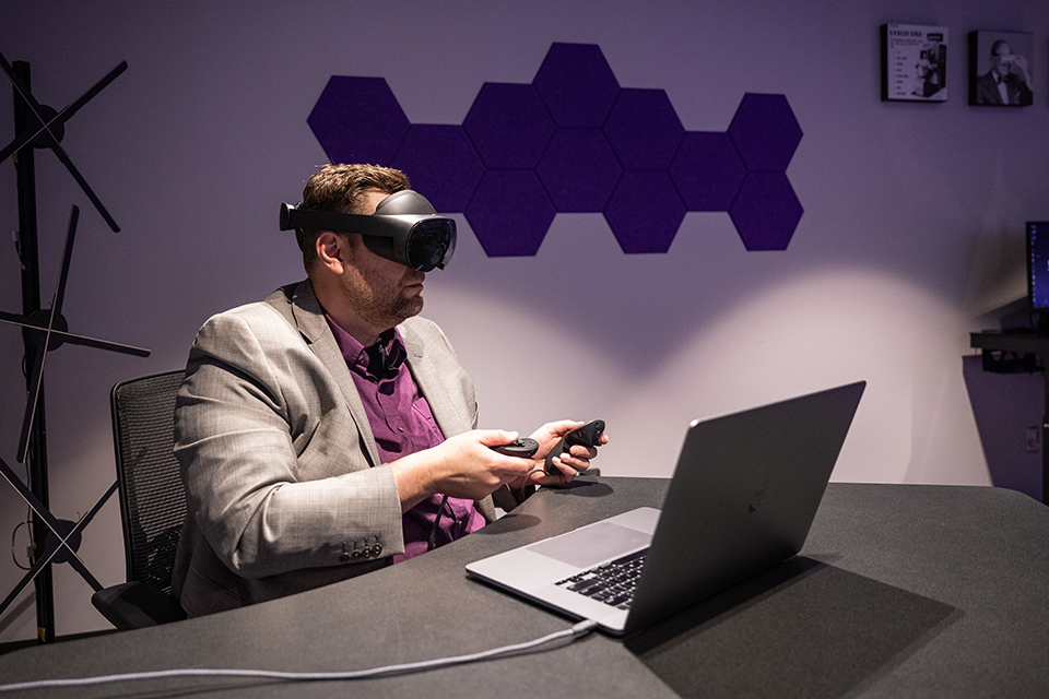 A professor in a gray sport jacket sits in a purple-lit room and at a desk, using a virtual reality headset to teach a class virtually.