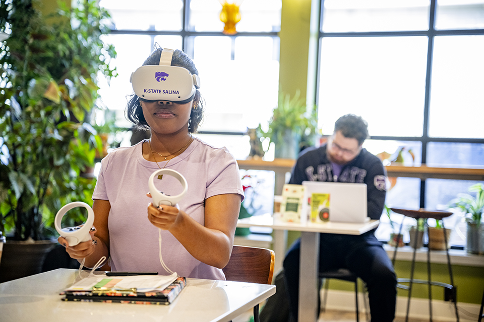 A student in a light lavender T-shirt sits at a table in a brightly lit cafe. She is wearing a virtual reality headset covering her eyes, and she's holding two white paddles with loops at their ends to manipulate the virtual environment she is experiencing.