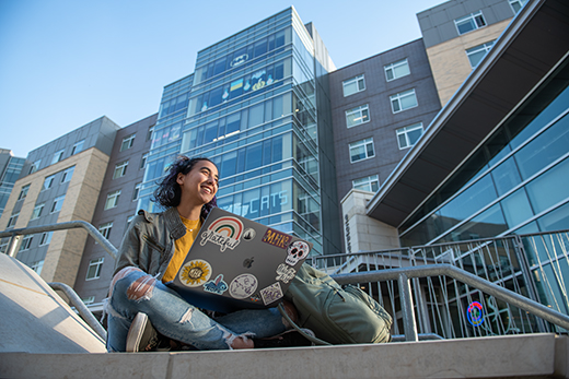 A female student sits cross-legged on a concrete stoop outside a tall, glass-paneled residence hall.