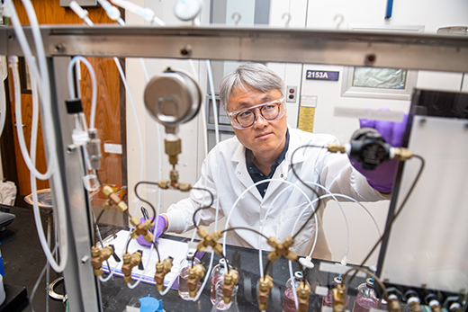 Wearing a white lab coat and safety goggles, Jeongdae Im sits behind and adjusts a row of hoses at a laboratory work bench.