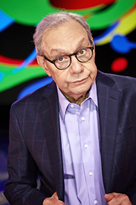 A man in a purple suit with his head titled down looks up at the camera through his glasses.