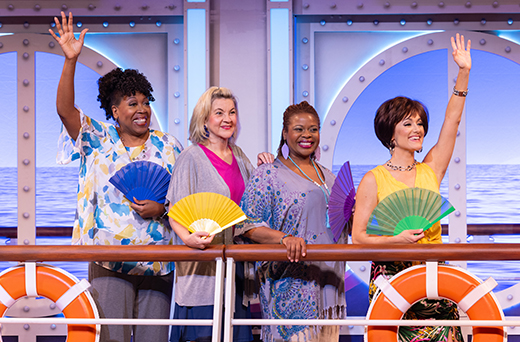 a scene from Menopause the Musical 2. Four women stand on a ship's deck holding colorful fans and waving, with life preservers and ocean-themed decor in the background.