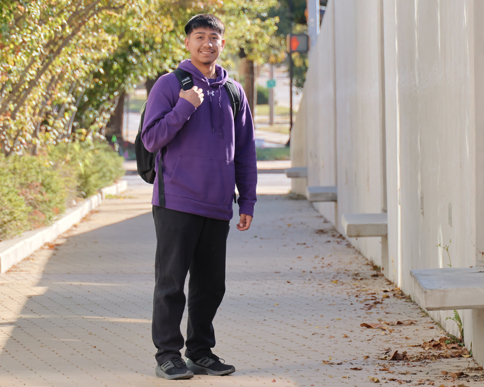 Wearing a purple hoodie and a black packpack, Bryant Tamayo stands at the end of a long walkway outside the Carl R. Ice College of Engineering Complex at K-State.