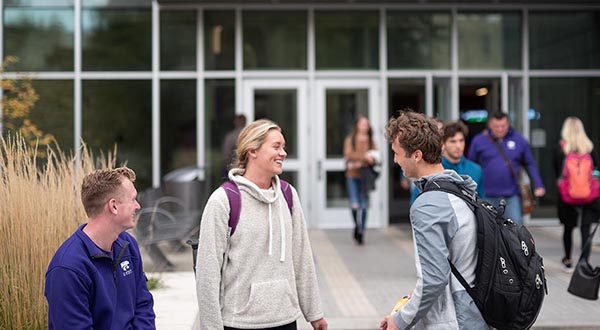 A group of students chat outdoors in front of a glass building at Kansas State University.