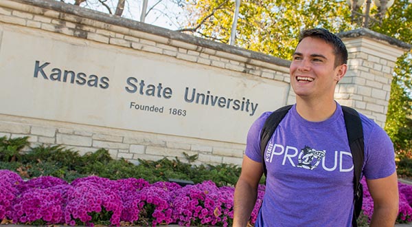 A male student wearing a purple shirt stands in front of one of Kansas State University's main entrance signs.