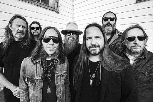 Black and white photo of the seven male musicians with facial hair in the country band Blackberry Smoke in dark clothing standing in front of a building with siding.