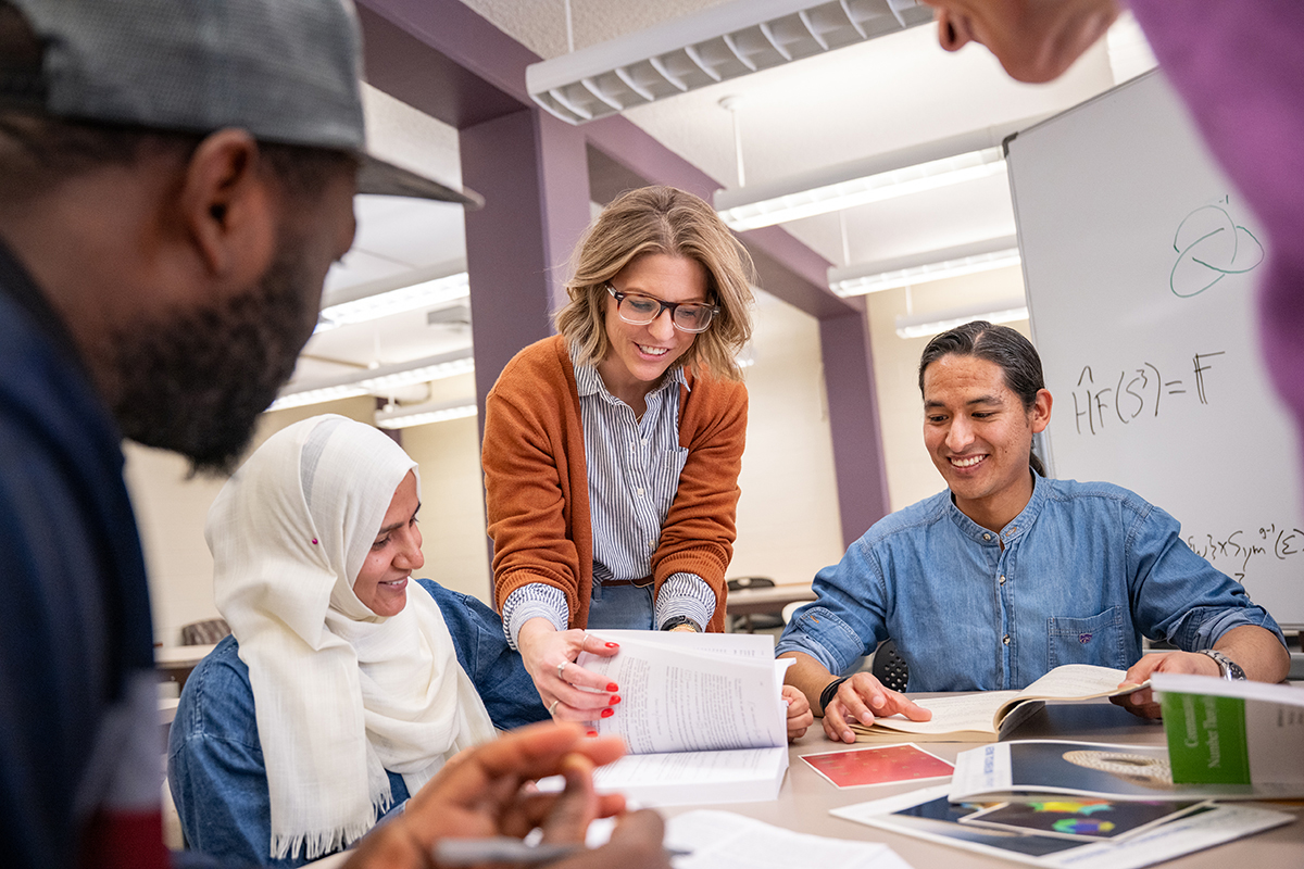 A female faculty memebr in a burnt orange blazer with a striped shirt and glasses directs students to a specifica page in a book.