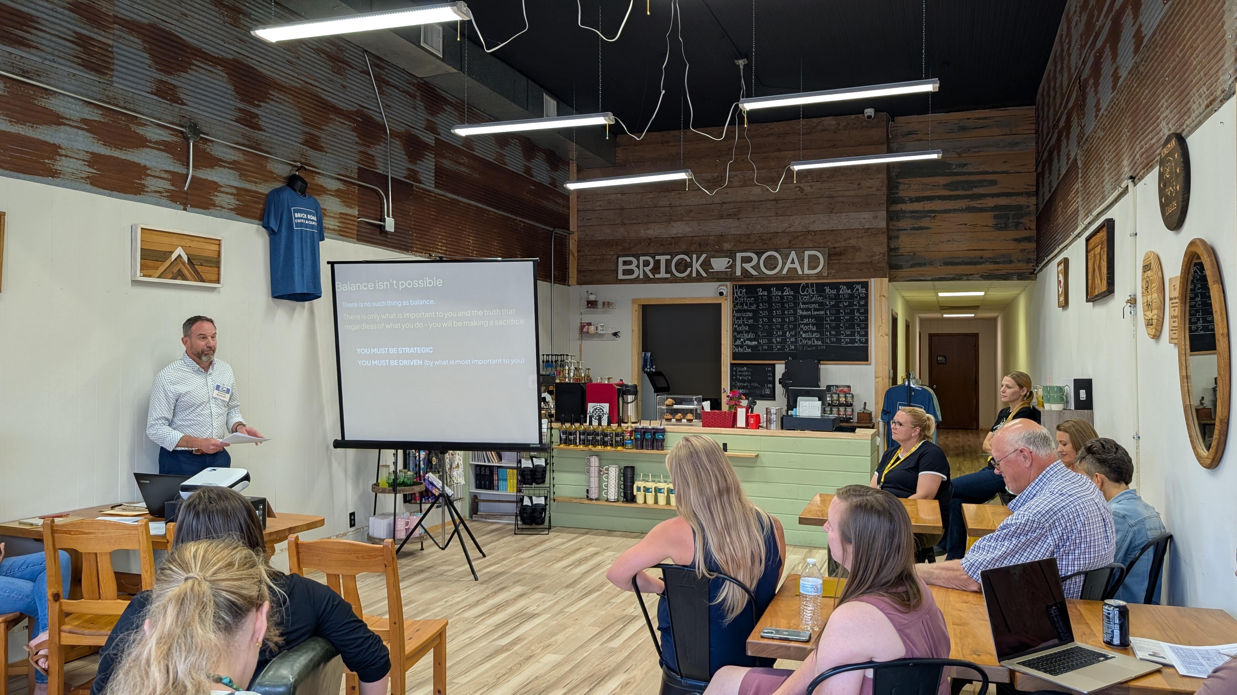A man in a button-up shirt presents to a room full of people with a projector screen at the front of the room at a coffeeshop called Brick Road.