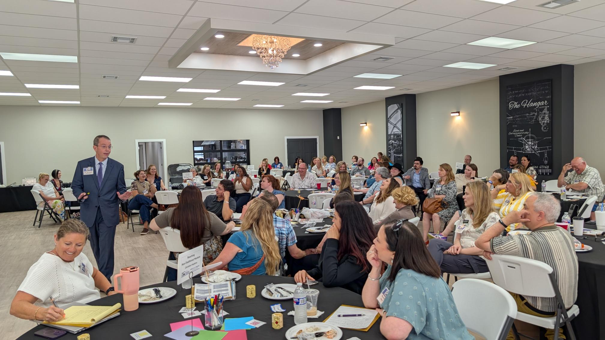 A man in a gray suit presents to a room full of people seated at round tables.