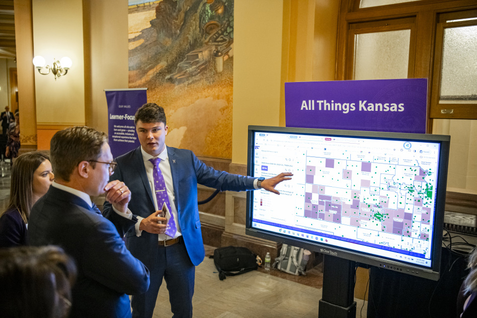 A student in a suit and purple tie gestures to a screen and talks to a group of people.