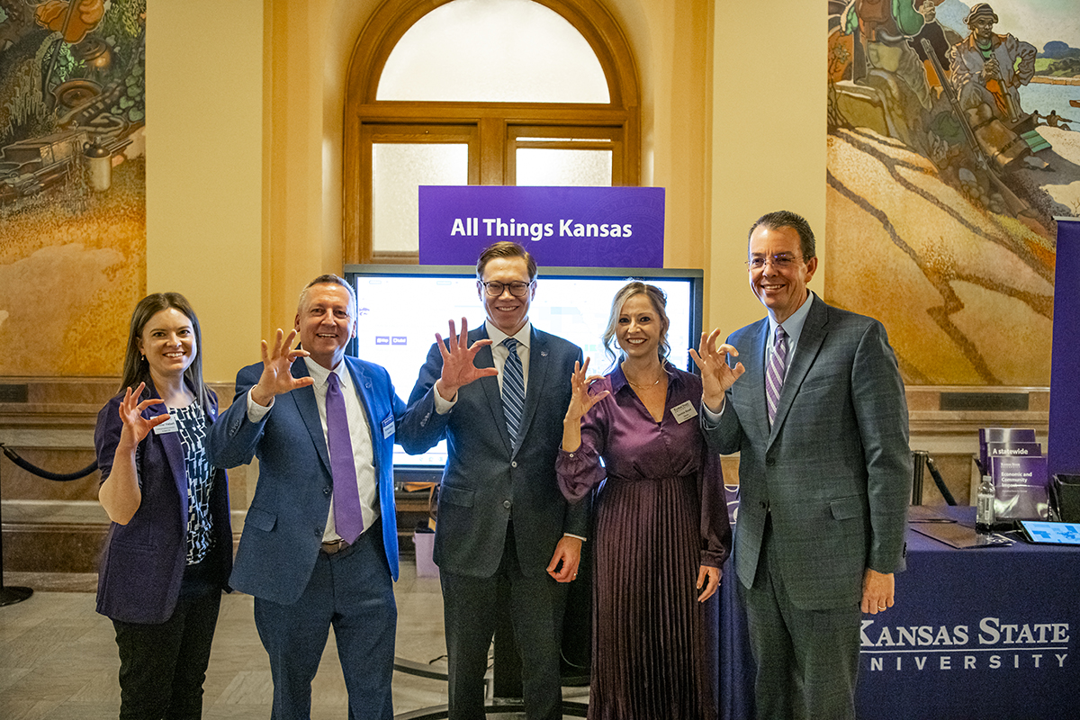 Five people in formal attire stand in front of a screen displaying All Things Kansas and hold up Wildcat hand symbols.
