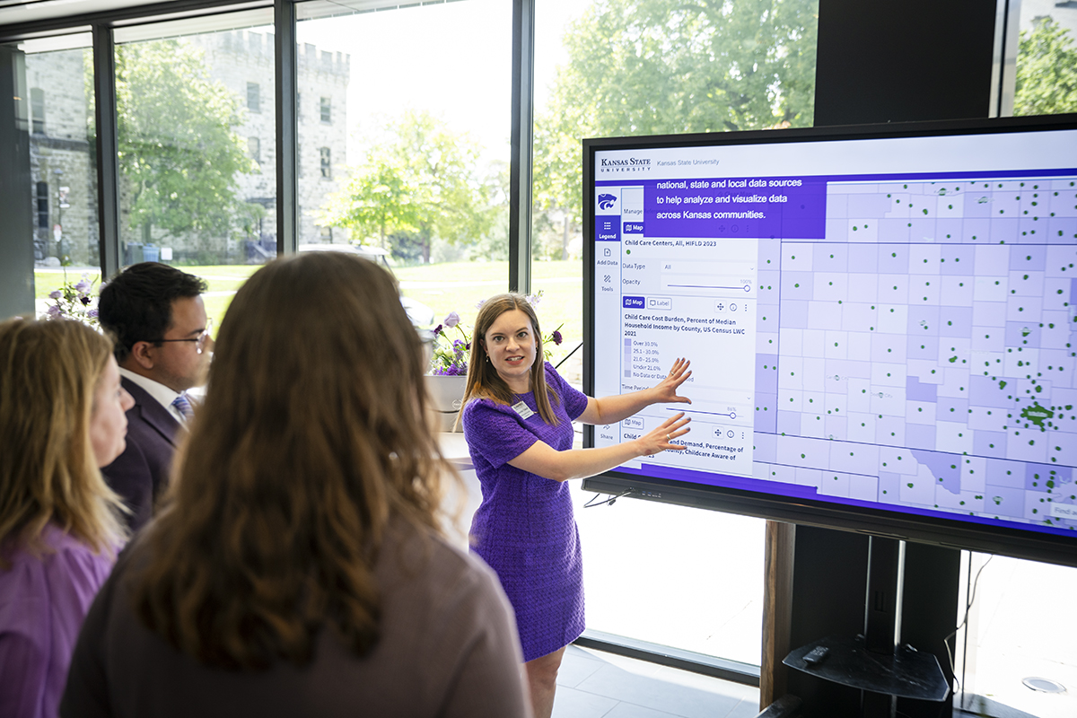 A woman in a purple dress talks to a group of people and gestures at a screen showing a map of Kansas.