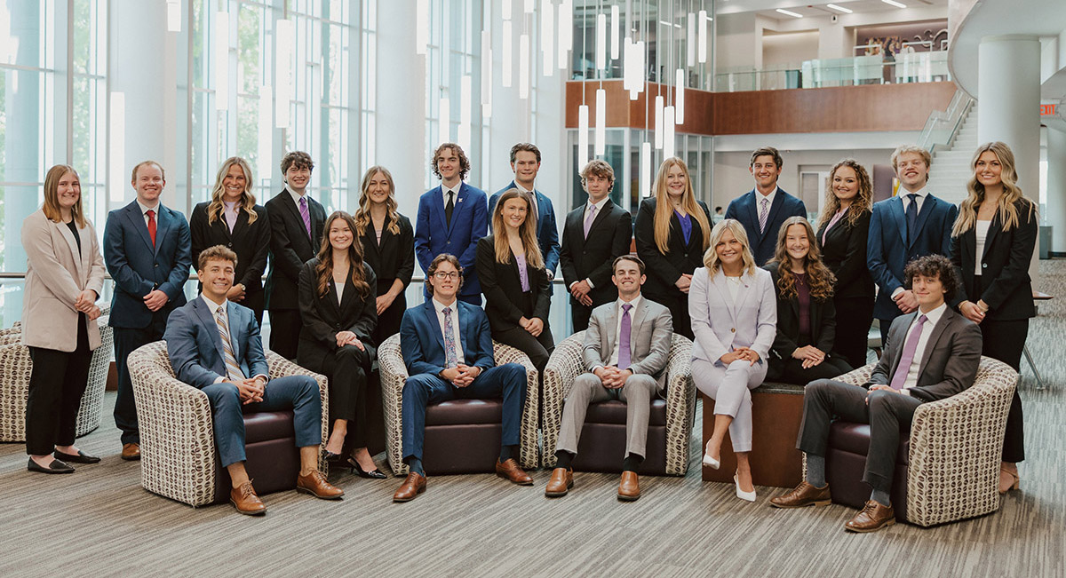 A large group of students, all in business professional attire, smile for a group photo. These are the Sales Ambassadors, and some sit in comfortable chairs while others stand, all forming a semicircle.