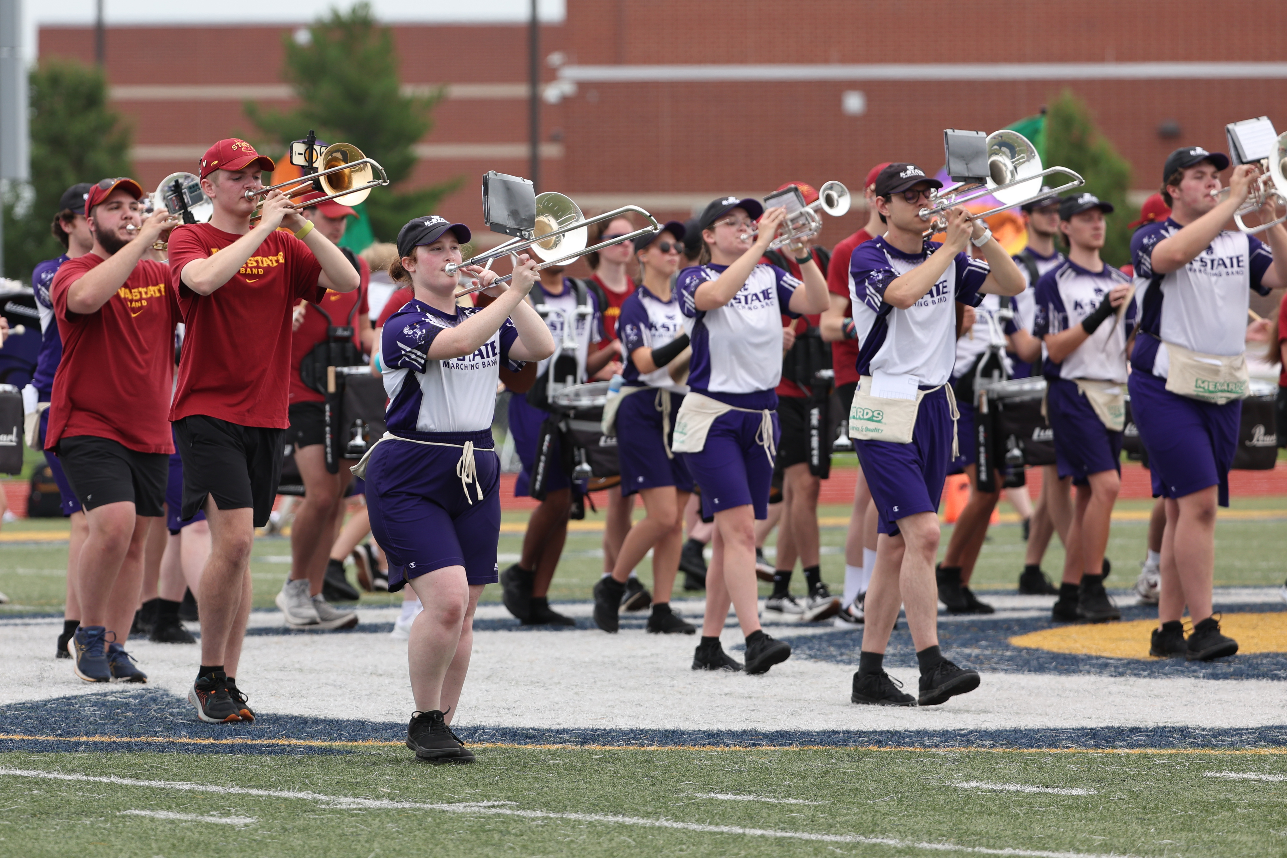 Wearing red and purple shirts alternatively, the combined corps of two college marching bands rehearse together on a football field.